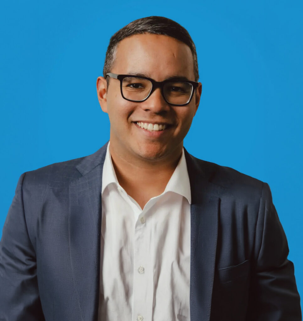 Smiling man in glasses and a dark blazer posed against a solid blue background.